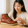 レッドウィングのオロラセットブーツに補色クリームを丁寧に塗り込む日本人女性が、明るい室内で革の手入れをしている様子を写した写真。ブーツケアに必要なブラシやクリームも自然に配置され、清潔感と温かみのある雰囲気が伝わる。
