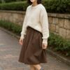 落ち着いた遊歩道で、クリーム色のニットとブラウンのミモレ丈スカートを着た日本人女性が、グレーの靴下を少しだけ見せる黒のショートブーツを合わせて立っている、明るく清潔感のあるコーディネート写真