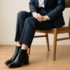 椅子に座った日本人女性がネイビーのスーツと黒のシンプルなショートブーツを着用し、清潔感のある室内で足元のマナー例として落ち着いた雰囲気で写っている様子。