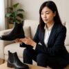 ビジネススーツを着た日本人女性が、明るく整った室内でサイドゴアブーツを手に取りながら丁寧に確認している様子を写した横長16:9の写真。