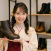 袴姿の日本人女性が靴店でレースアップの袴用ブーツを手に持って笑顔で立っている様子を写した写真。